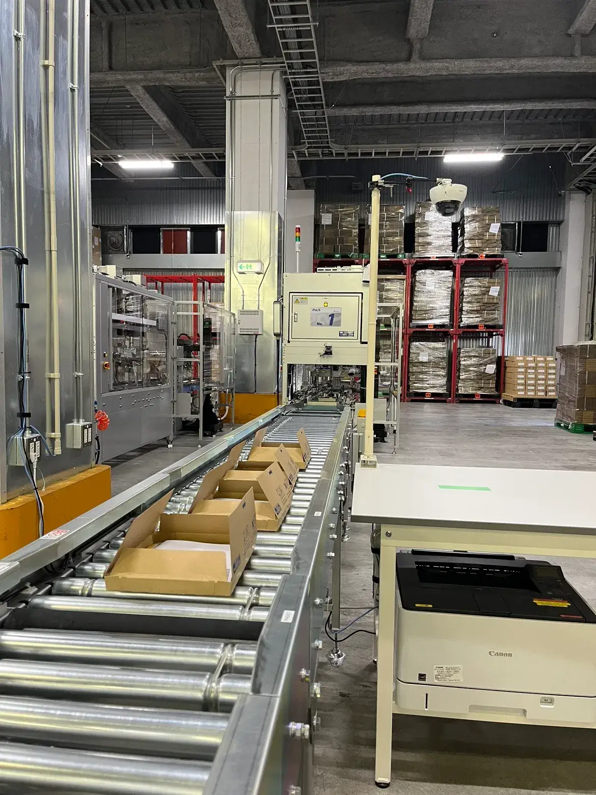 自動搬送ライン上に複数の段ボール箱が並び、物流倉庫内で梱包・仕分け作業が行われている様子。奥にはパレットに積まれた商品が保管され、右手前にはプリンターや制御機器が配置されている。