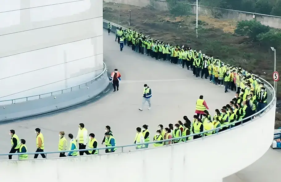 Large group of people wearing yellow safety vests walking in a line around a building, likely participating in an evacuation drill or safety inspection.