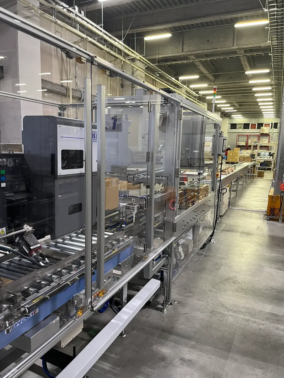 物流倉庫内の自動梱包ライン。透明カバーで囲まれた搬送装置の中を段ボール箱が流れ、奥では自動仕分けや検品作業が行われている。照明が整然と並ぶ広い工場空間で、効率的な物流システムを示している。