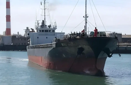 Cargo ship docked at a port, with a worker standing on the bow preparing for departure or arrival, and a factory with chimneys visible in the background.