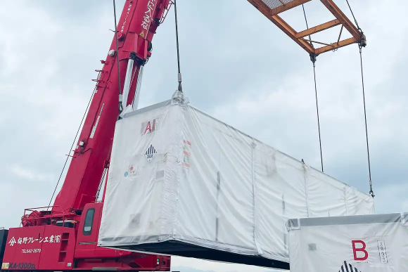 赤い大型クレーンで白い保護フィルムに包まれたコンテナを吊り上げて移動させている様子。