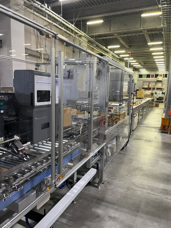 物流倉庫内の自動梱包ライン。透明カバーで囲まれた搬送装置の中を段ボール箱が流れ、奥では自動仕分けや検品作業が行われている。照明が整然と並ぶ広い工場空間で、効率的な物流システムを示している。