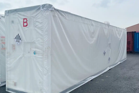 白いフィルムで全体を覆われた大型コンテナ。側面には危険物ラベルと識別用の「B」文字が表示され、屋外で保管されている様子。