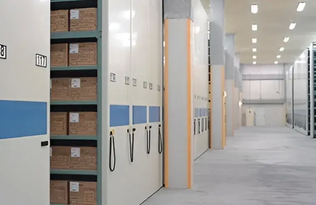 Cardboard boxes neatly stored on mobile shelves inside a warehouse, featuring white and blue panels with hand-crank mechanisms for moving the racks.