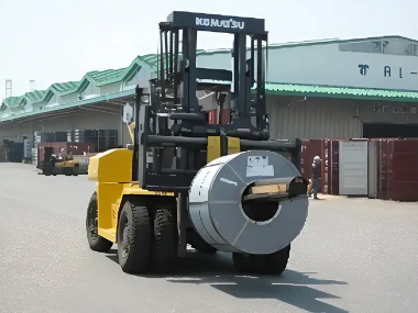 黄色いフォークリフトが金属コイルを運搬している様子。屋外の物流倉庫前で、重量物を慎重に取り扱う作業が行われている。