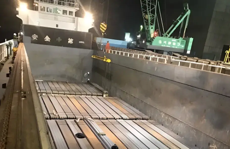 Nighttime port scene showing workers and a crane loading cargo onto a vessel, with metal sheets neatly arranged inside the ship’s hold.