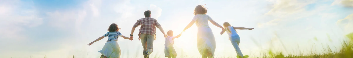 Panoramic view of a family holding hands and running forward across a grassy field under a bright, backlit sky.