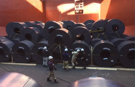 Multiple large steel coils stacked in a cargo hold, with a worker securing them with safety belts during pre-shipment loading for sea transport.