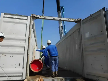 屋外でコンテナ内に赤いドラム缶を積み込む作業員たち。青い作業服と安全ヘルメットを着用し、クレーンを使って荷役作業を行っている様子。
