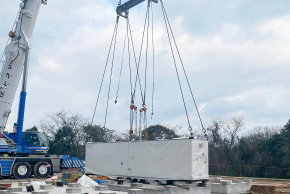 大型クレーンで白い保護フィルムに包まれたコンテナを吊り上げ、基礎ブロック上に設置している様子。