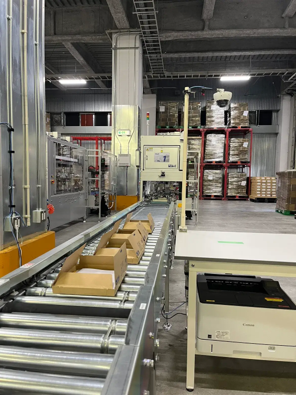 自動搬送ライン上に複数の段ボール箱が並び、物流倉庫内で梱包・仕分け作業が行われている様子。奥にはパレットに積まれた商品が保管され、右手前にはプリンターや制御機器が配置されている。